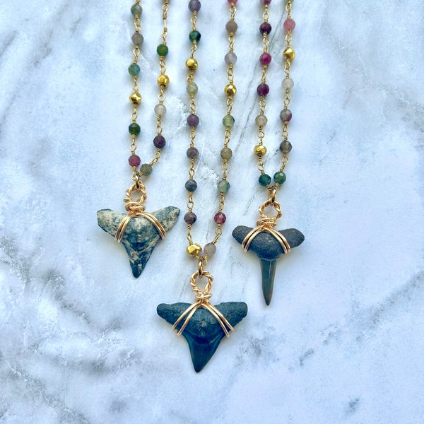 May include: Three necklaces with shark tooth pendants. The pendants are wrapped in gold wire and suspended from strands of colorful beads. The beads are a mix of green, purple, and gold. The necklaces are displayed on a white marble surface.