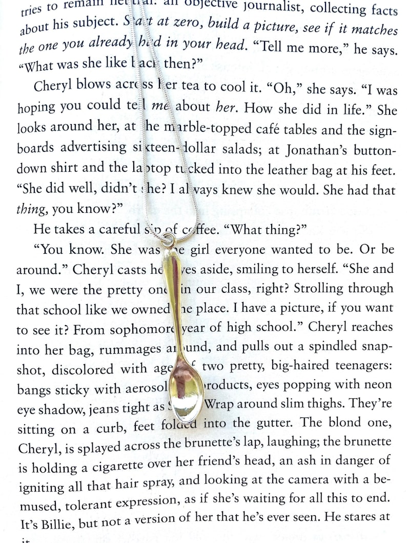 May include: A close-up of a gold-colored metal spoon with a white handle. The spoon is partially obscured by a book page with text about a young woman named Cheryl. The text describes Cheryl as a popular girl in high school.