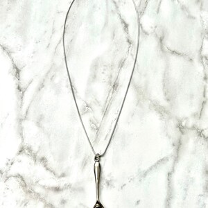 May include: A silver spoon pendant necklace on a silver chain. The spoon is hanging from the chain and is facing downwards.
