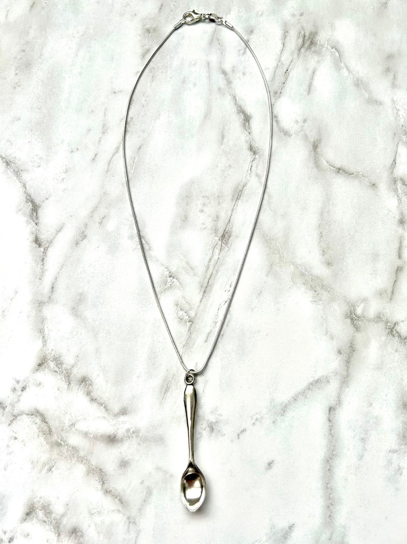May include: A silver spoon pendant necklace on a silver chain. The spoon is hanging from the chain and is facing downwards.
