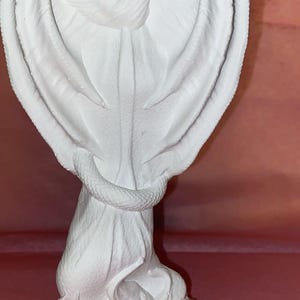 Puede incluir: Una escultura blanca de una criatura alada con cuernos, posiblemente un drag&oacute;n o un demonio. La figura tiene grandes alas detalladas y una serpiente enrollada alrededor de su cuerpo. La escultura est&aacute; sobre una base texturizada.