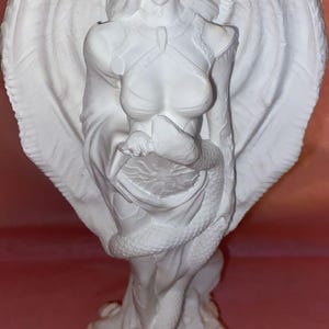 Puede incluir: Una escultura blanca de una figura alada con cuernos, una serpiente y un vestido fluido. La figura tiene una expresi&oacute;n serena y est&aacute; de pie sobre una base detallada. Las alas est&aacute;n extendidas y la serpiente est&aacute; enrollada alrededor de la figura.