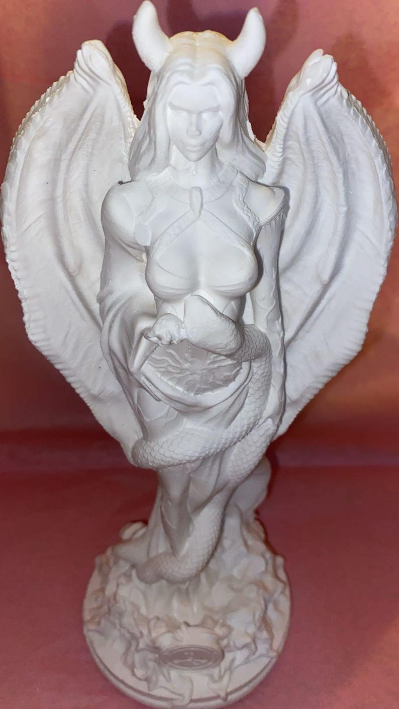 Puede incluir: Una escultura blanca de una figura alada con cuernos, una serpiente y un vestido fluido. La figura tiene una expresi&oacute;n serena y est&aacute; de pie sobre una base detallada. Las alas est&aacute;n extendidas y la serpiente est&aacute; enrollada alrededor de la figura.