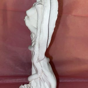 Puede incluir: Escultura blanca de una mujer con una capucha de cobra y una serpiente enrollada a su alrededor. La estatua est&aacute; sobre una base texturizada, y el fondo es de color rosa liso.