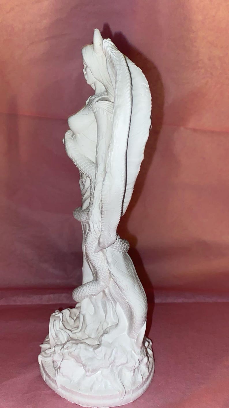 Puede incluir: Escultura blanca de una mujer con una capucha de cobra y una serpiente enrollada a su alrededor. La estatua est&aacute; sobre una base texturizada, y el fondo es de color rosa liso.