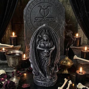 Puede incluir: Una estatua gris oscuro de Lilith sosteniendo una calavera, con una tableta de piedra detr&aacute;s. La estatua est&aacute; rodeada de velas, huesos y p&eacute;talos de rosa secos. La palabra "Lilith" est&aacute; inscrita en la base de la estatua.