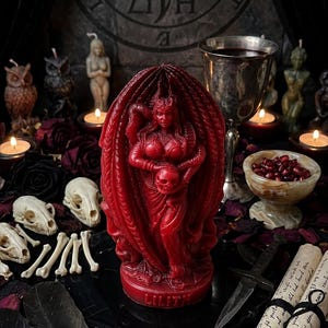 Puede incluir: Una vela roja de Lilith, una figura que sostiene una calavera, es el punto focal de esta imagen. La vela est&aacute; rodeada de calaveras, huesos, plumas y pergaminos. Tambi&eacute;n hay otras velas y un c&aacute;liz, creando un tema g&oacute;tico.