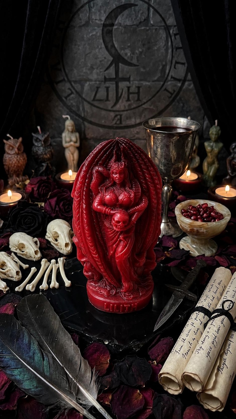 Puede incluir: Una vela roja de Lilith, una figura que sostiene una calavera, es el punto focal de esta imagen. La vela est&aacute; rodeada de calaveras, huesos, plumas y pergaminos. Tambi&eacute;n hay otras velas y un c&aacute;liz, creando un tema g&oacute;tico.