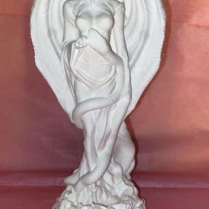 Puede incluir: Una escultura blanca de una figura femenina con grandes alas y cuernos, envuelta en un vestido fluido. Una serpiente est&aacute; enrollada alrededor de la figura. La escultura est&aacute; sobre un pedestal con un s&iacute;mbolo.
