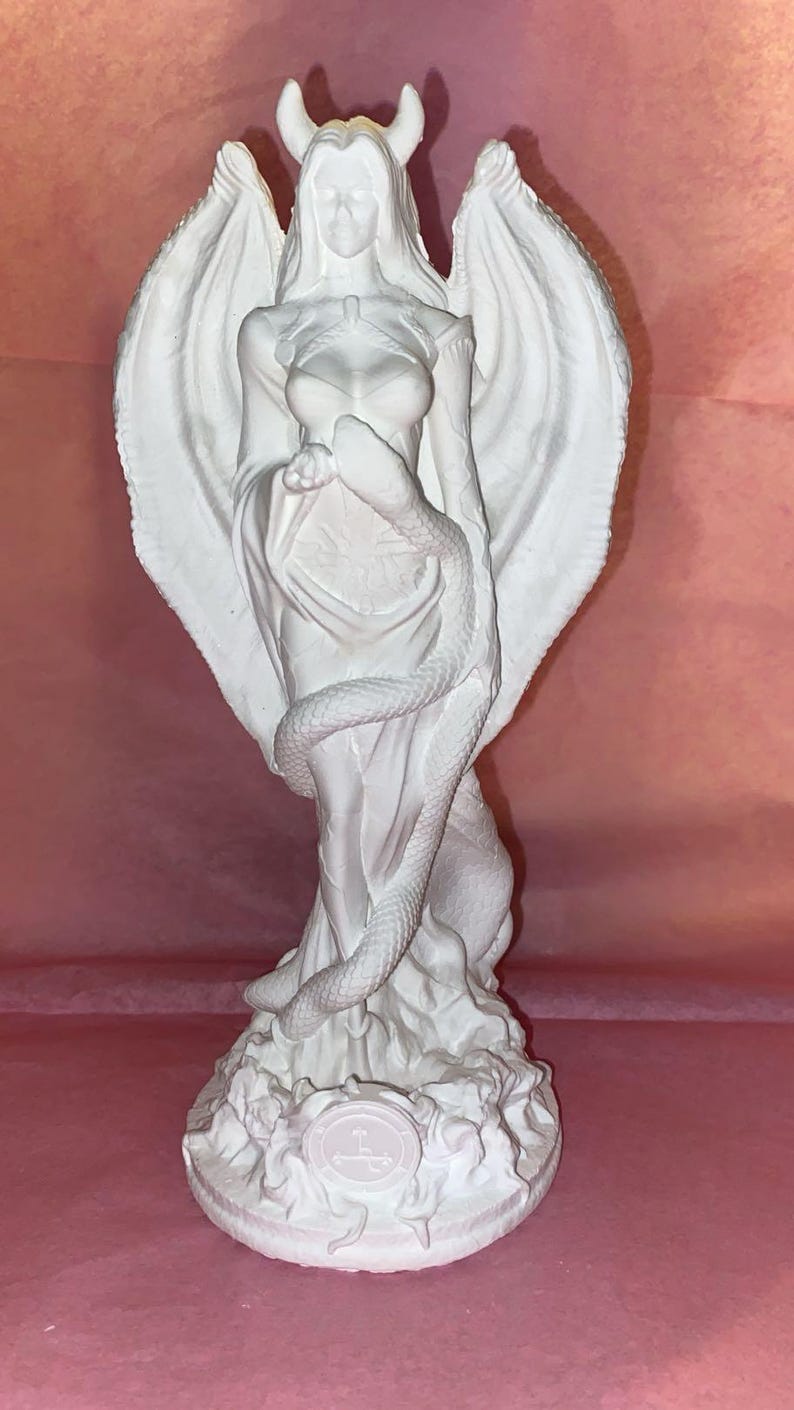 Puede incluir: Una escultura blanca de una figura femenina con grandes alas y cuernos, envuelta en un vestido fluido. Una serpiente est&aacute; enrollada alrededor de la figura. La escultura est&aacute; sobre un pedestal con un s&iacute;mbolo.