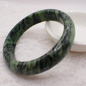 May include: A green and black jade bangle bracelet. The smooth, polished bracelet has a marbled appearance with varying shades of green and black. The bracelet is circular and sits on a white surface.