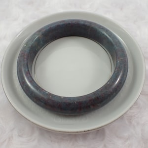 May include: A dark blue and gray jade bangle bracelet with red and white flecks. The bangle is resting on a white saucer.