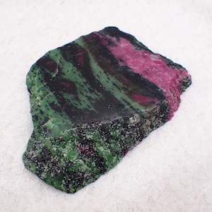 May include: A rough-cut stone featuring a striking combination of colors. The stone displays a deep green hue with black veining, contrasted by a vibrant magenta section. The bottom edge has a speckled, granular texture.