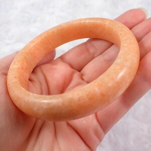 May include: A peach-colored, polished stone bangle bracelet. The bracelet is a solid, circular band with a smooth, glossy finish. The color is a warm, light orange with subtle variations in tone.