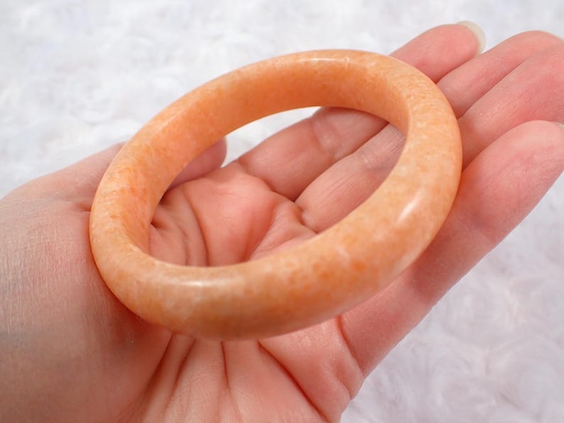 May include: A peach-colored, polished stone bangle bracelet. The bracelet is a solid, circular band with a smooth, glossy finish. The color is a warm, light orange with subtle variations in tone.