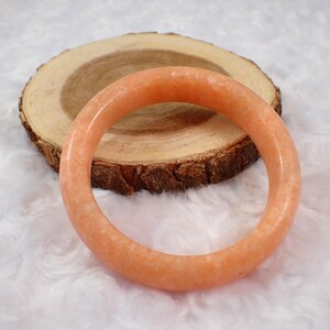May include: A peach-colored, round bangle bracelet. The bracelet is made of a smooth, polished material and is resting on a wooden slice with a natural bark edge. The background is a soft, white, textured surface.