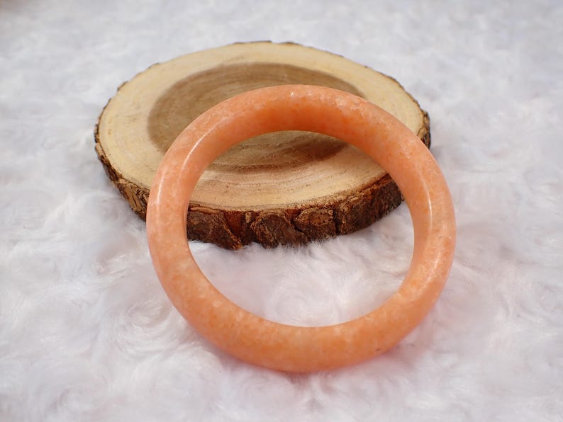 May include: A peach-colored, round bangle bracelet. The bracelet is made of a smooth, polished material and is resting on a wooden slice with a natural bark edge. The background is a soft, white, textured surface.