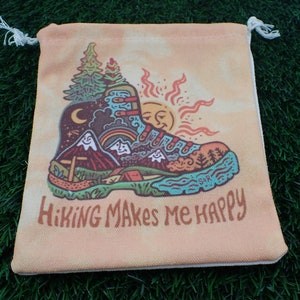 May include: A drawstring bag with a yellow, orange, and blue tie-dye background. The bag features a cartoon illustration of a hiking boot with a mountain scene, a sun, and a moon. The text "Hiking Makes Me Happy" is printed in black below the boot.