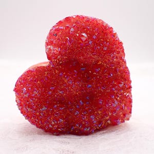 May include: A heart-shaped, red crystal formation with a sparkling, textured surface. The crystal has a vibrant, iridescent quality, with hints of purple and gold. The object is set against a soft, white background.