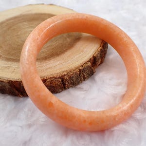 May include: An orange-colored gemstone bracelet. The bracelet is circular and sits on a wooden slice with a white, fluffy background. The bracelet's color is a light, translucent orange.