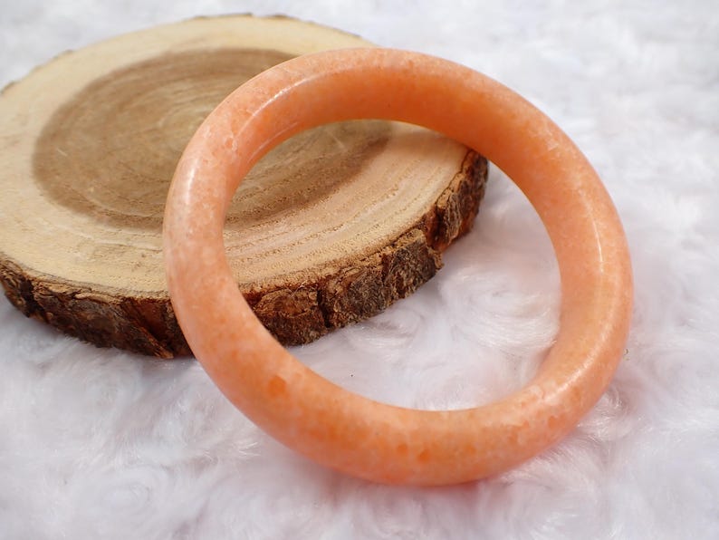 May include: An orange-colored gemstone bracelet. The bracelet is circular and sits on a wooden slice with a white, fluffy background. The bracelet's color is a light, translucent orange.
