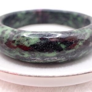 May include: A faceted ring made from a stone with green, black, and red colours. The ring is set on a white surface. This gemstone ring is a unique piece of jewellery.