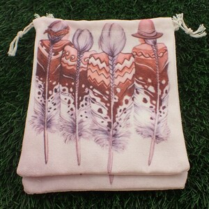 May include: A pink drawstring bag with a white canvas base. The bag features a design of four women with feathers in their hair. The women are wearing colorful clothing and hats.