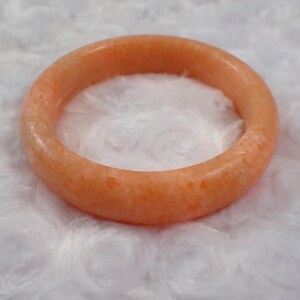 May include: A round, orange-colored bangle bracelet. The bracelet is made of a solid, opaque material with a slightly textured surface. The jewelry is set against a white, fluffy background.