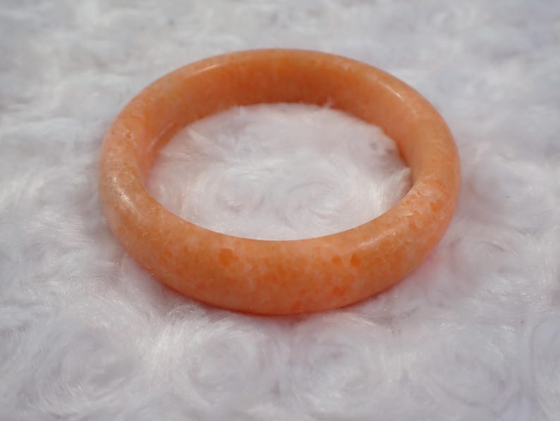 May include: A round, orange-colored bangle bracelet. The bracelet is made of a solid, opaque material with a slightly textured surface. The jewelry is set against a white, fluffy background.