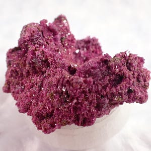 May include: A close-up of a vibrant, textured, magenta-colored mineral specimen. The mineral has a porous, coral-like appearance with small, sparkling crystals. The specimen is set against a soft, white background.