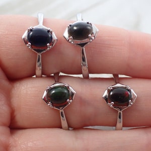May include: Four silver rings with oval-shaped gemstones. The rings have a decorative design around the stones. The stones are black, dark green, and dark red.