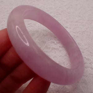 May include: A light purple jade bangle bracelet. The smooth, polished bracelet is a soft lavender color. The bangle is round and appears to be made of a solid piece of stone. The background is a white, textured surface.