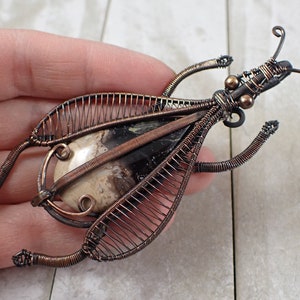 May include: A copper wire insect brooch with a brown and white agate stone. The wings are made of wire and the body is detailed with wire curls.