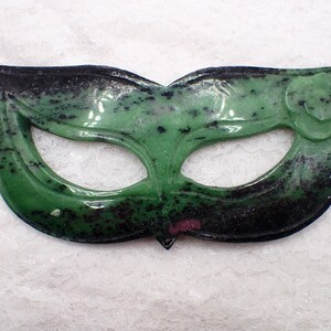 May include: A green and black gemstone masquerade mask with a floral accent. The mask has eye cutouts and a curved shape. The gemstone has a glossy finish and a speckled pattern. The mask is likely for costume or decorative purposes.