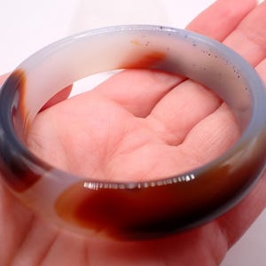 May include: A translucent agate bangle bracelet. The bracelet features a blend of colors, including white, gray, brown, and red. The smooth, polished surface of the bracelet reflects light, highlighting its natural patterns and variations. A piece of jewelry.