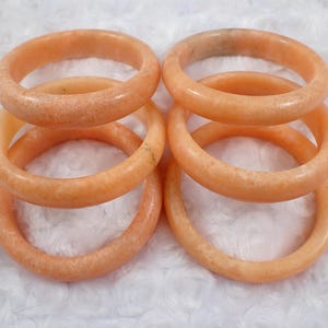 May include: Six peach-colored stone bangles are stacked and arranged on a white, textured surface. The bracelets have a smooth, polished finish and a speckled appearance. The bangles are a warm, neutral color.