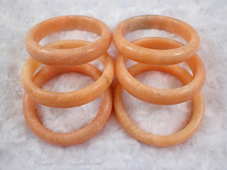 May include: Six peach-colored stone bangles are stacked and arranged on a white, textured surface. The bracelets have a smooth, polished finish and a speckled appearance. The bangles are a warm, neutral color.