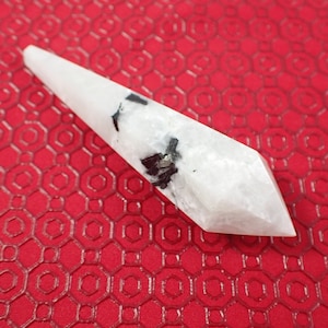 May include: A white crystal point with black inclusions, also known as a selenite wand, on a red background.
