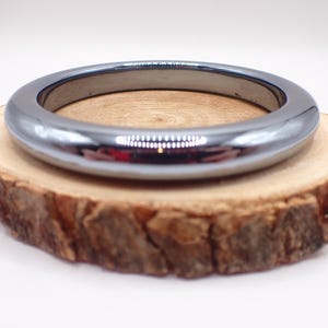 May include: A polished, dark gray bangle bracelet with a smooth, rounded design. The bracelet is displayed on a rustic, circular wooden slice with a natural bark edge. The background is a soft white.