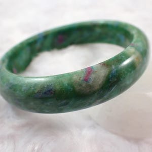 Könnte beinhalten: Ein grünes Jade-Armreif-Armband mit roten und blauen Einschlüssen. Die glatte, polierte Oberfläche des Armbands reflektiert das Licht und hebt die natürlichen Variationen des Steins hervor. Das Armband hat eine kreisförmige Form und liegt auf einer weißen Oberfläche.