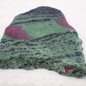 May include: A rough-cut slab of zoisite with ruby inclusions. The stone features a vibrant green base color with black streaks and patches of deep red ruby. The edges are uneven, showcasing the natural, raw form of the mineral specimen.