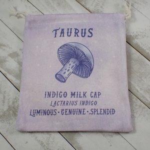 May include: A light purple drawstring bag with the word "TAURUS" printed in white at the top. A purple and white illustration of a mushroom is in the center of the bag. The text "INDIGO MILK CAP" is printed below the mushroom, followed by "LACTARIUS INDIGO" and "LUMINOUS - GENUINE - SPLENDID".