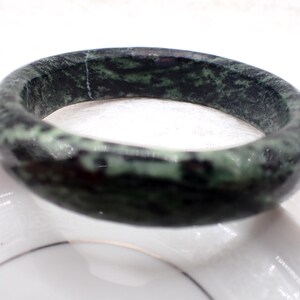 May include: A circular jade bracelet, displaying a mottled pattern of dark green and black. The polished stone bracelet is set against a white backdrop, highlighting its natural colours and textures. This is a classic piece of jewellery.