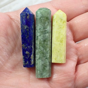 May include: Three crystal wands, one blue, one green and one yellow, are displayed on a hand. The wands are smooth and polished.