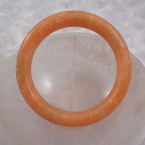 May include: A round, orange-colored bracelet. The bracelet is made of a solid material with a slightly textured surface. The bracelet is resting on a white, rounded surface.