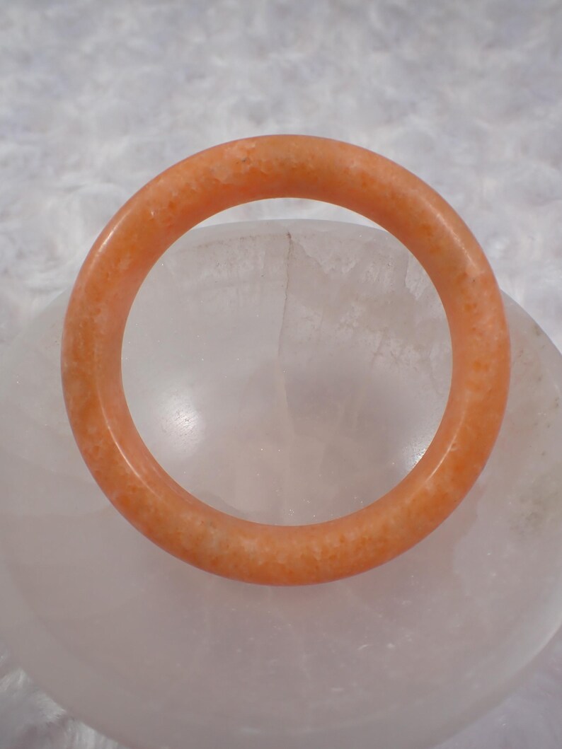 May include: A round, orange-colored bracelet. The bracelet is made of a solid material with a slightly textured surface. The bracelet is resting on a white, rounded surface.