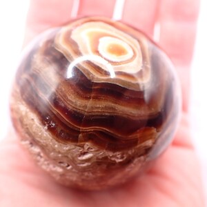 May include: A polished, spherical stone with a swirling pattern of brown, tan, and cream-colored bands. The stone has a smooth, glossy surface and a natural, earthy appearance. The stone is held in a hand.