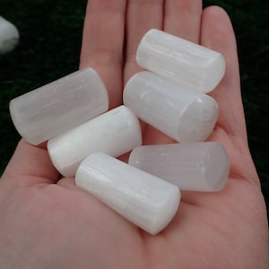 May include: A collection of seven white selenite sticks, also known as selenite wands, are displayed in a hand. The sticks are smooth and polished, with a slightly translucent appearance.