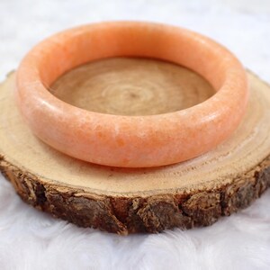 May include: A peach-colored, round gemstone bangle bracelet rests on a rustic wooden slice. The bracelet has a smooth, polished surface and a warm, inviting hue. The wood slice has a natural, textured appearance with visible grain.