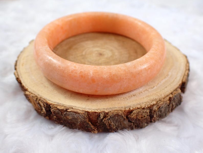 May include: A peach-colored, round gemstone bangle bracelet rests on a rustic wooden slice. The bracelet has a smooth, polished surface and a warm, inviting hue. The wood slice has a natural, textured appearance with visible grain.